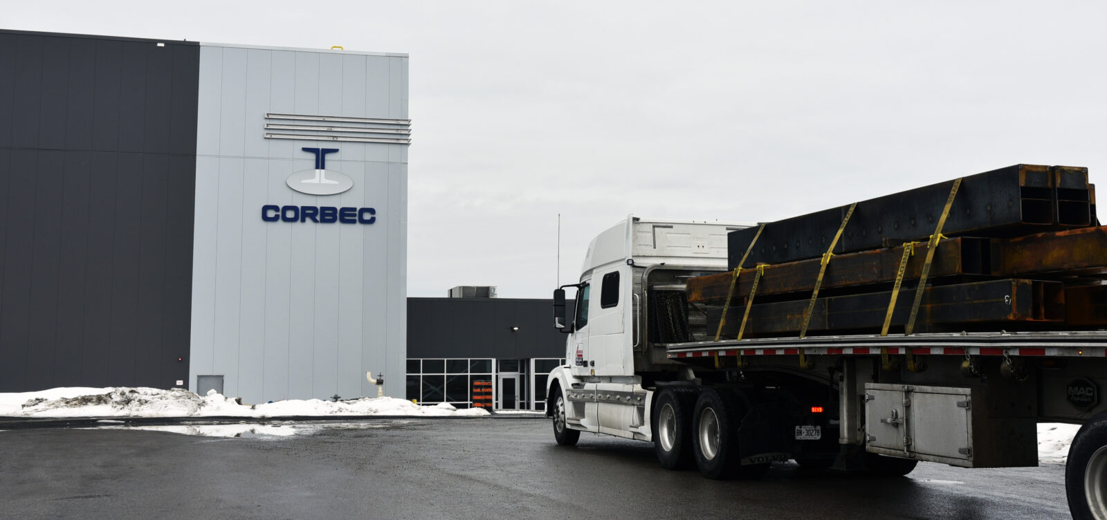 Exterior photo of the plant showing a double-height building with grey cladding and a prominent Corbec logo. A flatbed truck carrying rods of steel is in the foreground approaching the building.